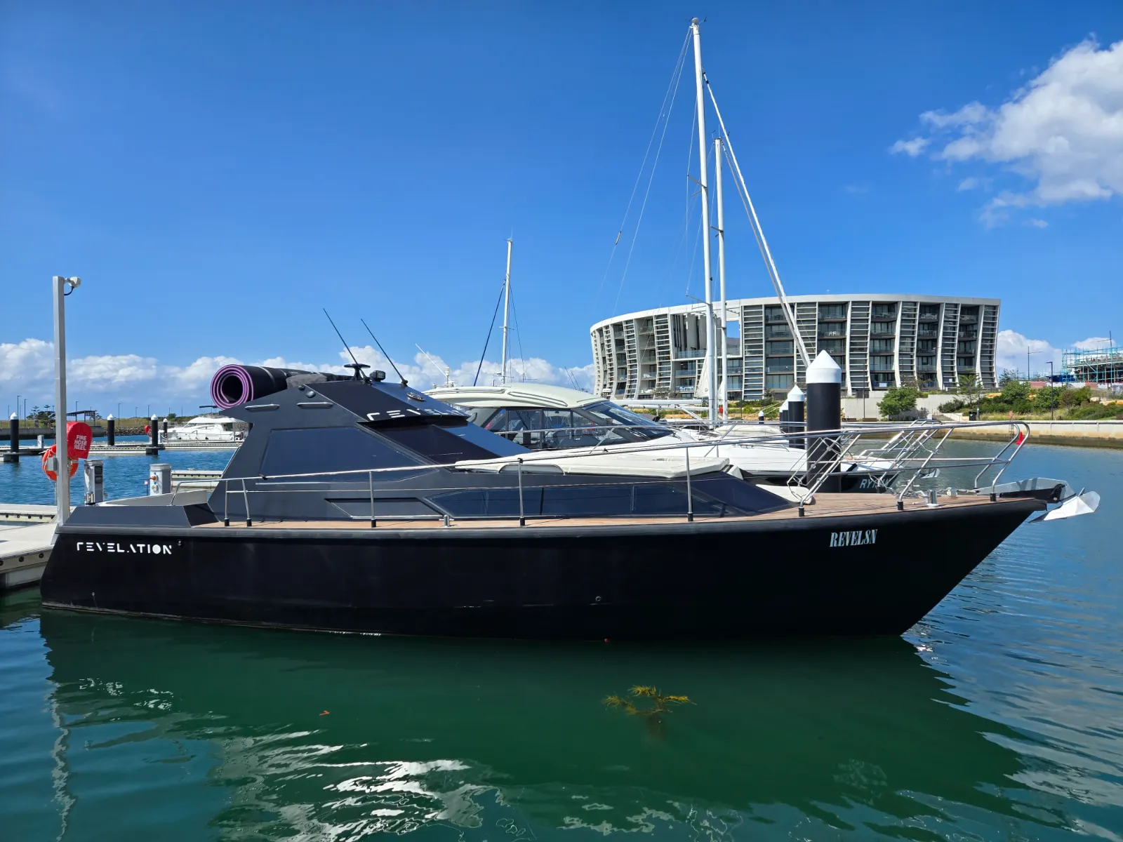 Revelation full profile from dock — satin black hull, teak decking, stadium behind