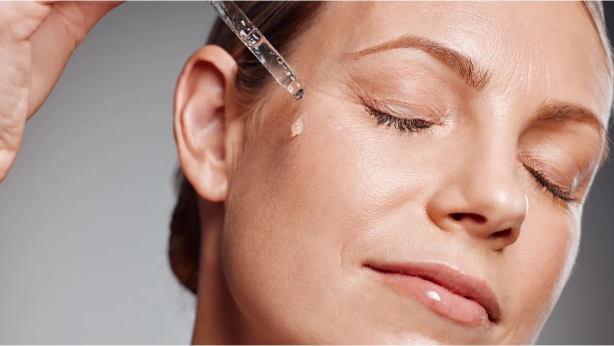 Woman applying a clear facial serum with a glass dropper to her cheekbone, eyes serenely closed