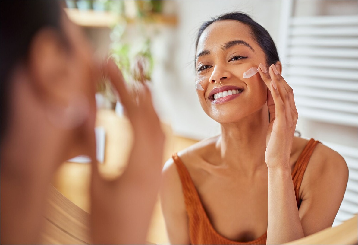 Smiling woman applying moisturizer to her cheek in a bright bathroom, reflecting a consistent daily skincare routine.