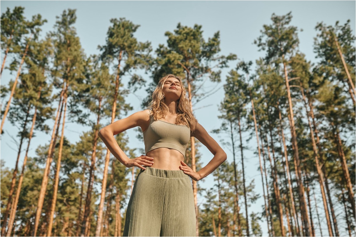 Woman standing outdoors among tall pine trees, face turned toward the sun.