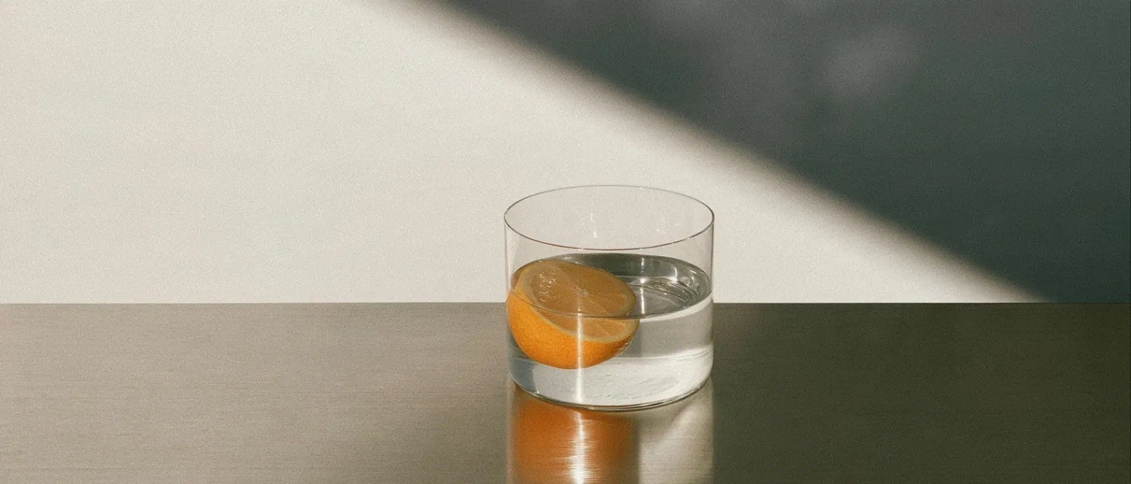 A short clear glass of still water with a halved lemon floating in it, standing on a dark bronze table in a hard diagonal of daylight