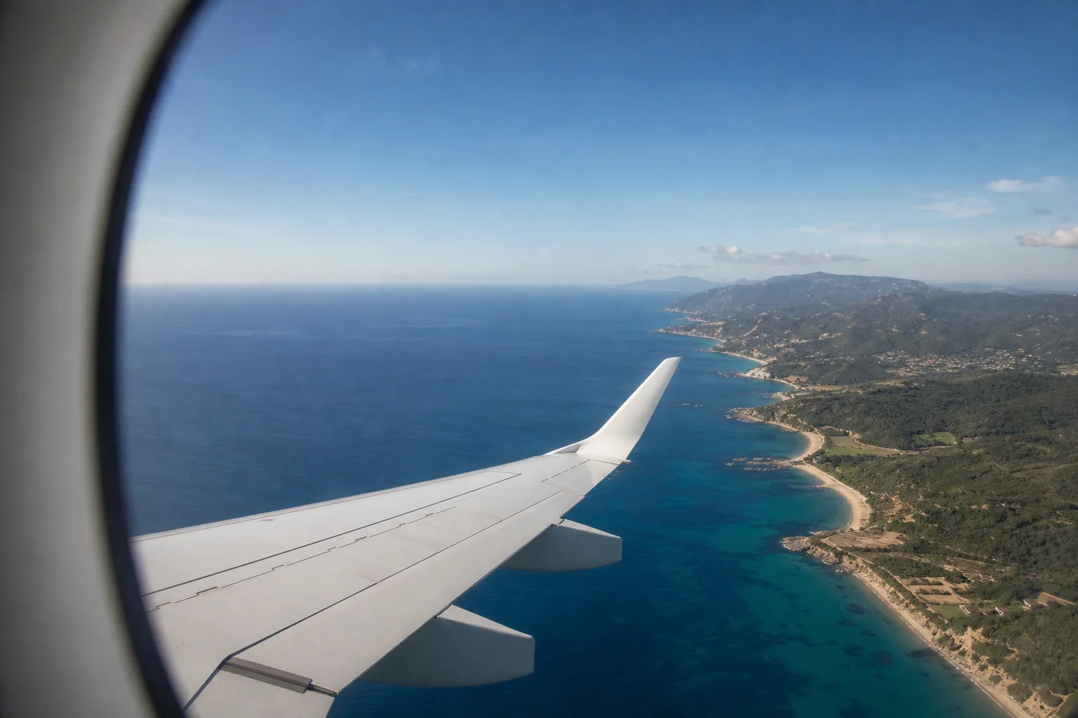 Airplane window view of a sunlit coastline and turquoise sea